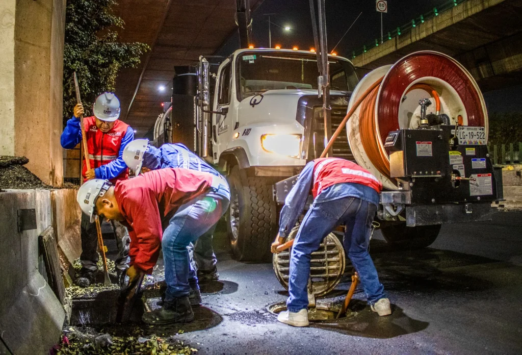 Operarios con casco trabajando de noche en una alcantarilla abierta junto a un camión de limpieza de redes de saneamiento
